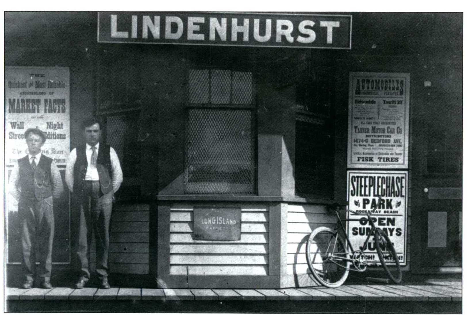 Two men in black and white photo standing in front of Lindenhurst train station. 